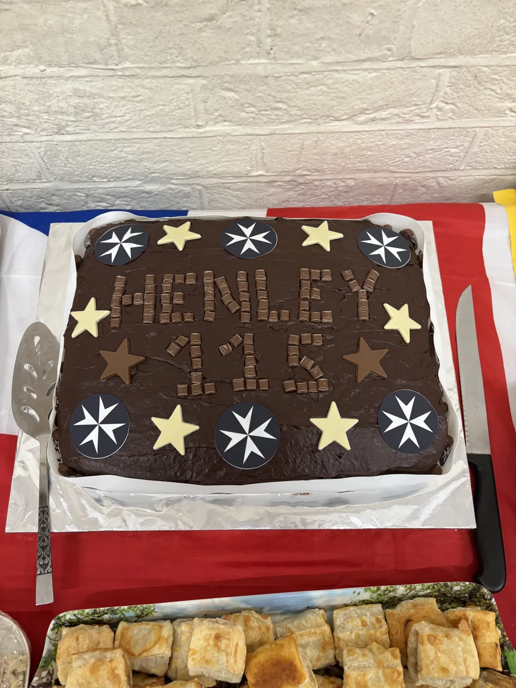 A rectangular chocolate cake with Henley 115 spelled out in chocolate pieces, decorated with white and dark chocolate stars and the St John Ambulance eight-pointed cross emblem, on a table covered with a Union flag